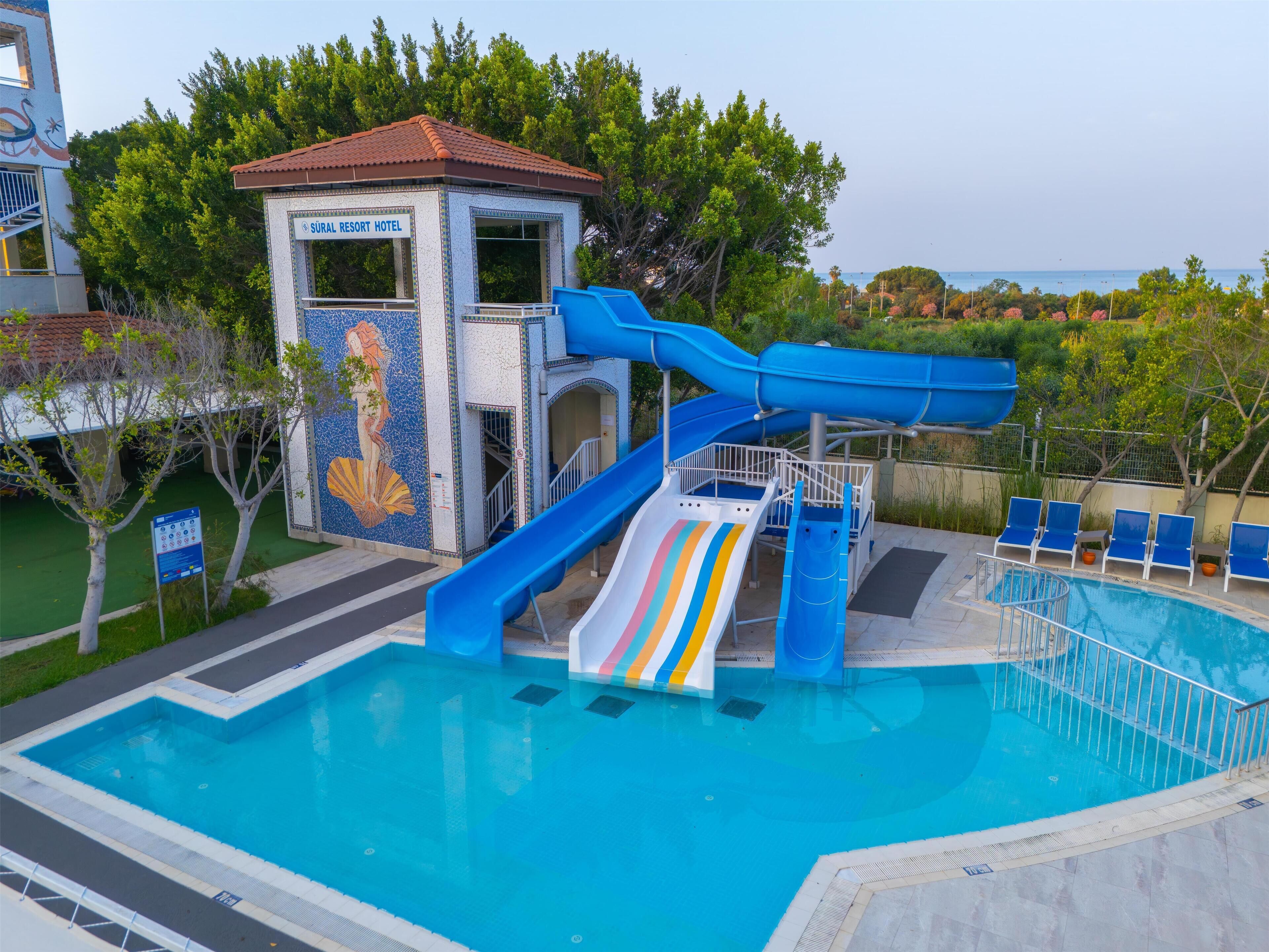 A swimming pool with a water slide, a small building labeled 'SURAL RESORT HOTEL', and a view of the surrounding greenery.