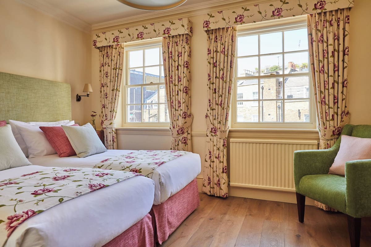 Une chambre avec deux lits, des rideaux à motifs floraux, un fauteuil vert et une vue sur les bâtiments à l’extérieur.