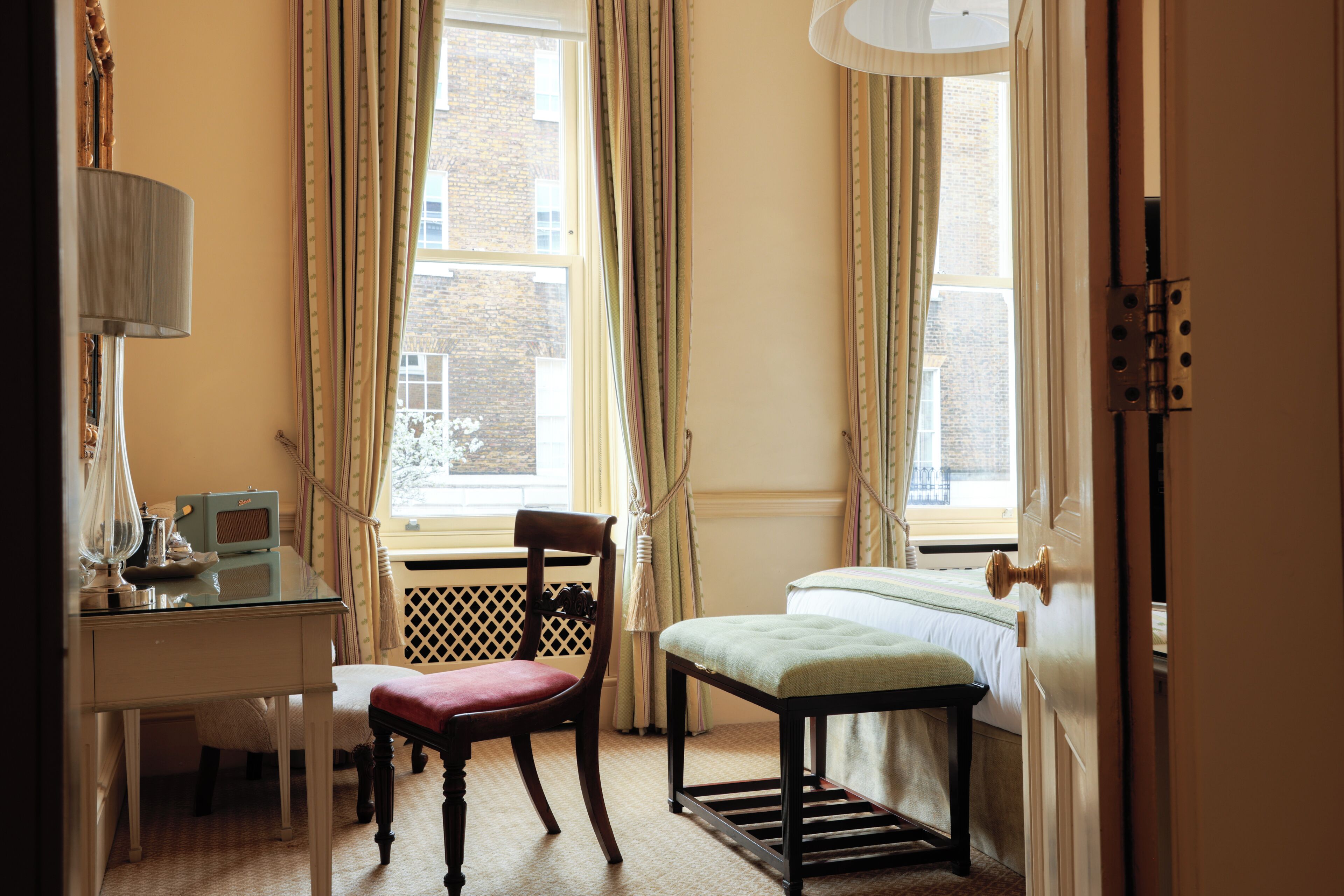 Ein Zimmer mit Schreibtisch, Stuhl und Bett, mit einem Fenster mit Vorhängen und Blick auf Gebäude.