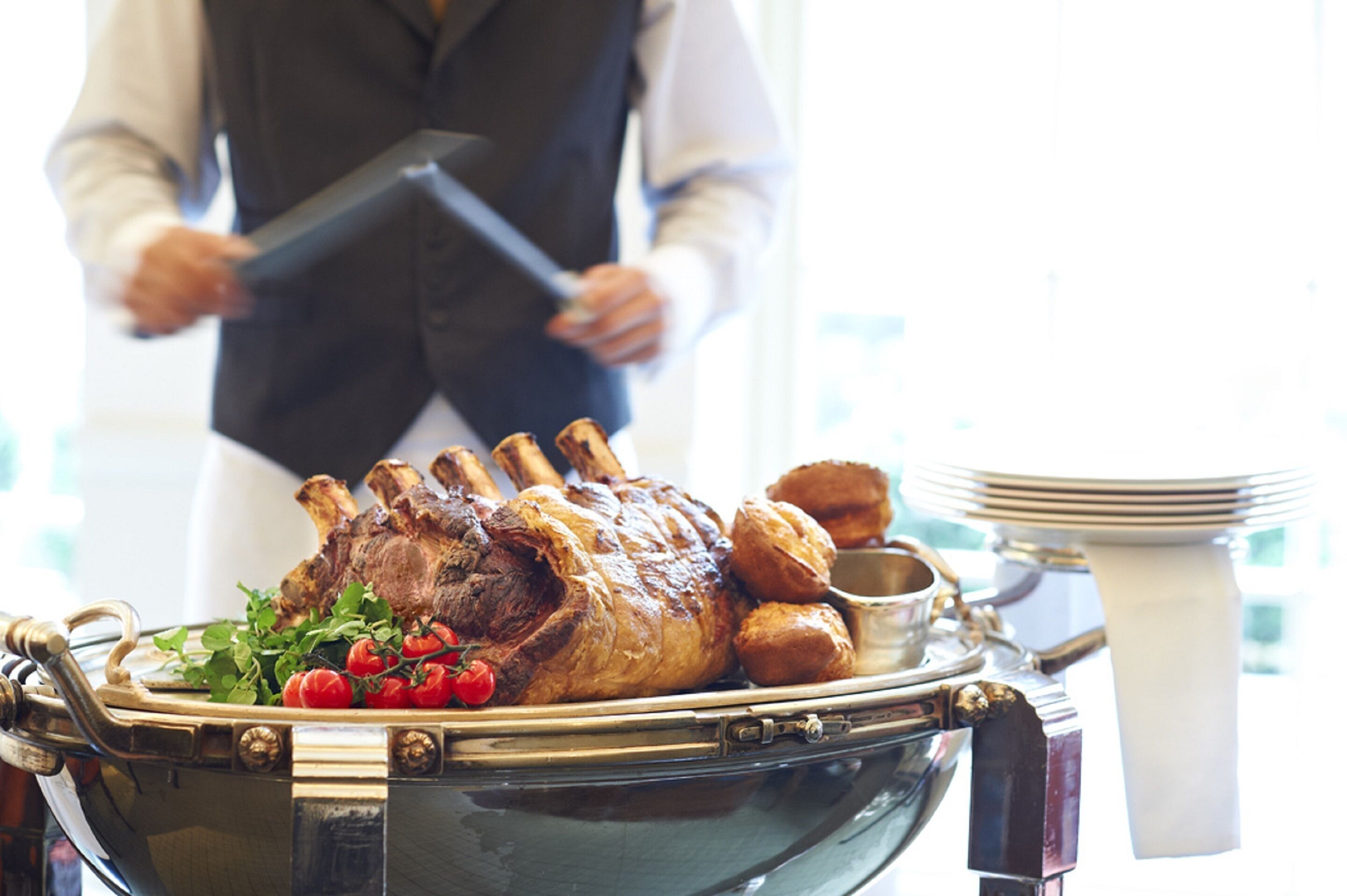 A roasted lamb leg on a silver platter with a garnish of herbs and tomatoes, accompanied by a serving of Yorkshire pudding and a small bowl of sauce, set on a table with a white napkin and a stack of plates.