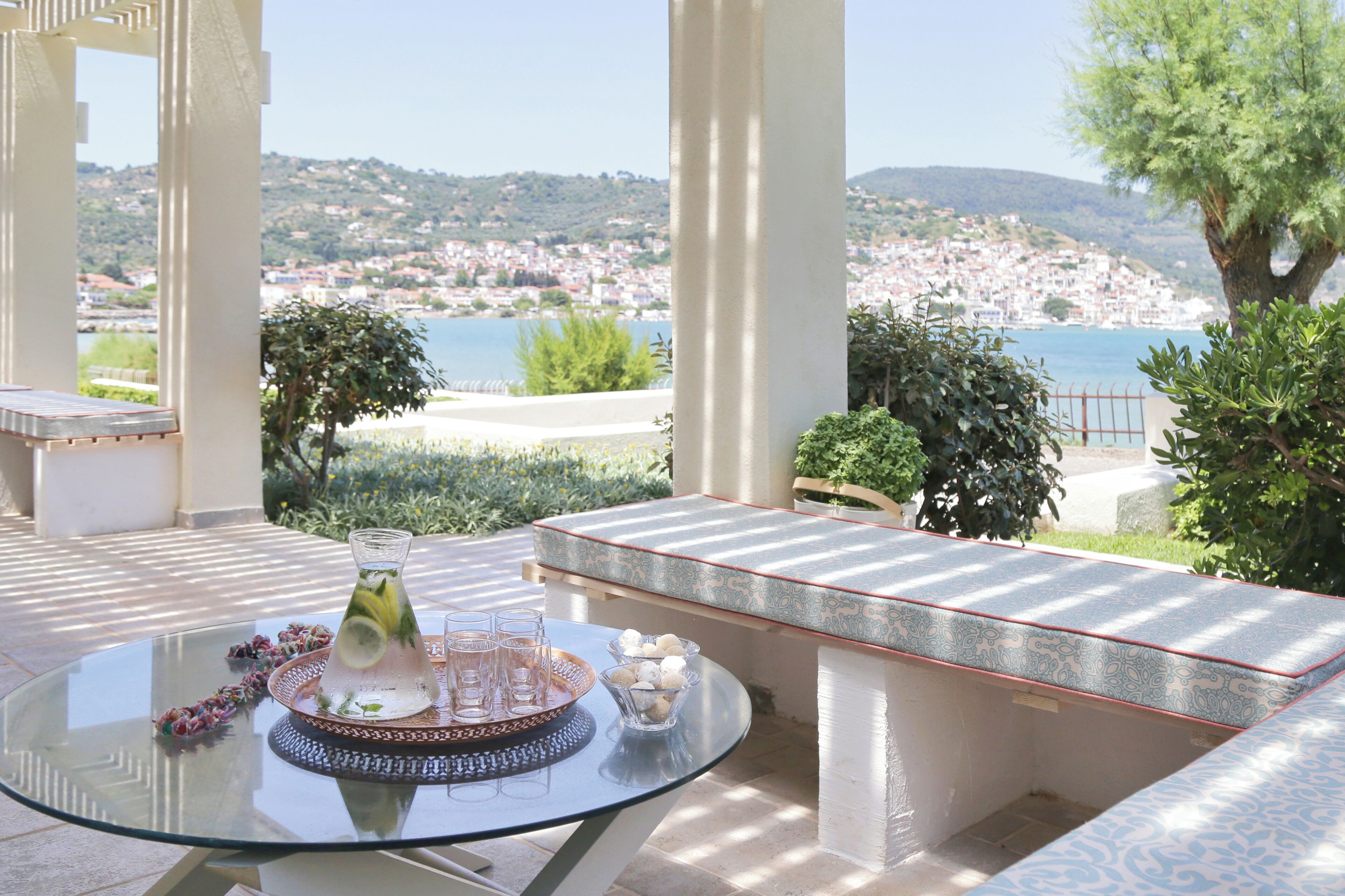 A glass table with a drink and glasses, a bench, and a view of a coastal town.