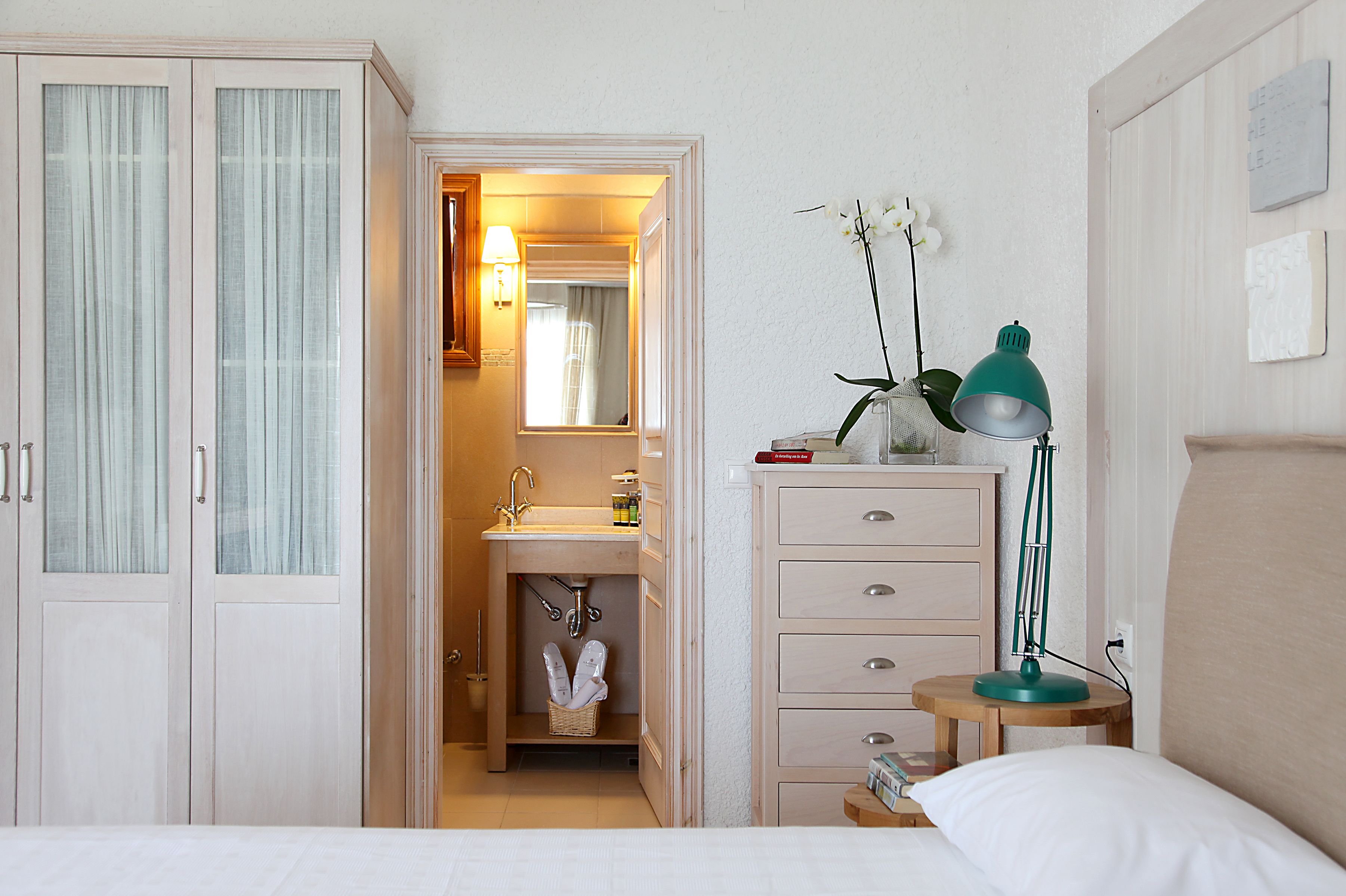 A hotel room with a bed, a bedside table with a lamp, a dresser, a vanity with a mirror, and a bathroom with a sink and towel rack.