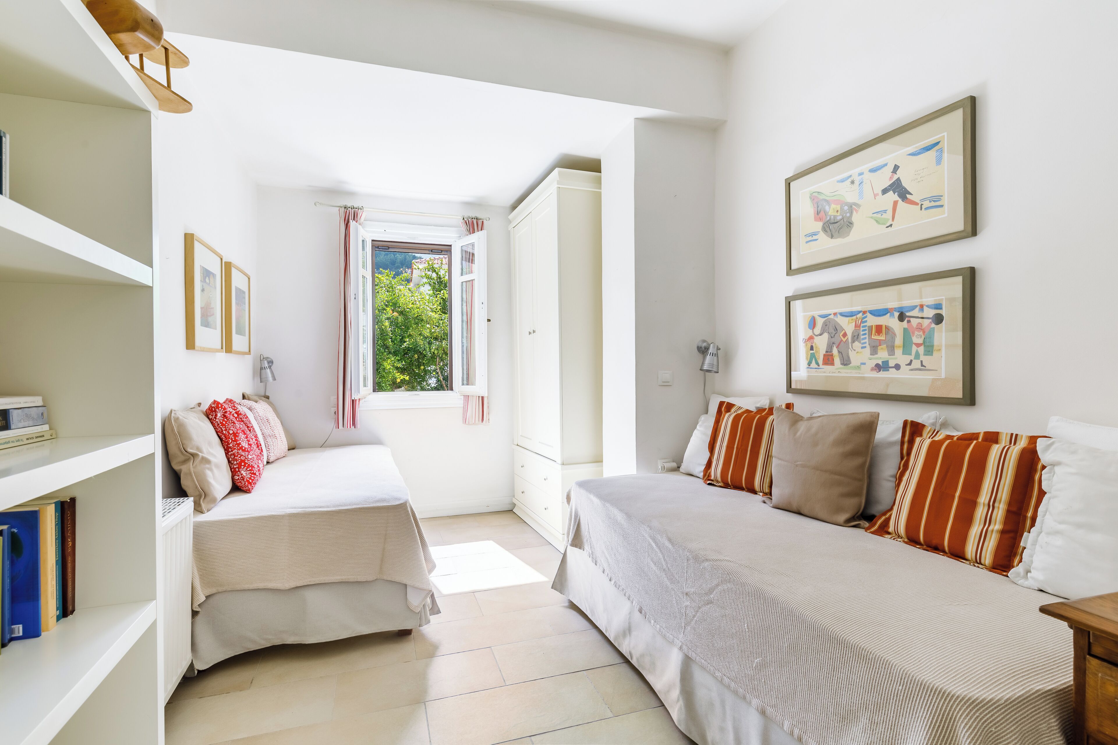 A bedroom with two beds, a window with curtains, a bookshelf, and framed artwork on the wall.