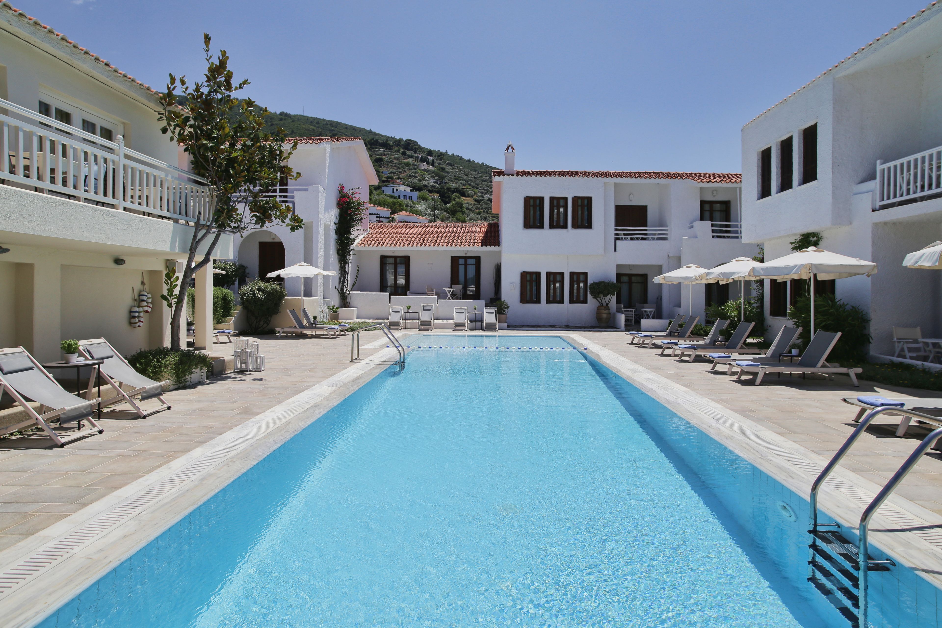 A swimming pool with lounge chairs and umbrellas, surrounded by buildings with balconies and a mountainous backdrop.