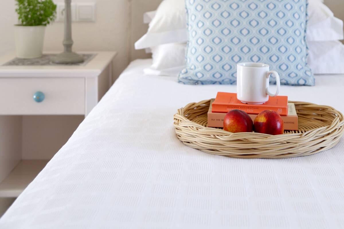 Una cama bien hecha con un edredón blanco, una almohada estampada en azul y una bandeja de mimbre con manzanas y una taza en una mesita de noche.