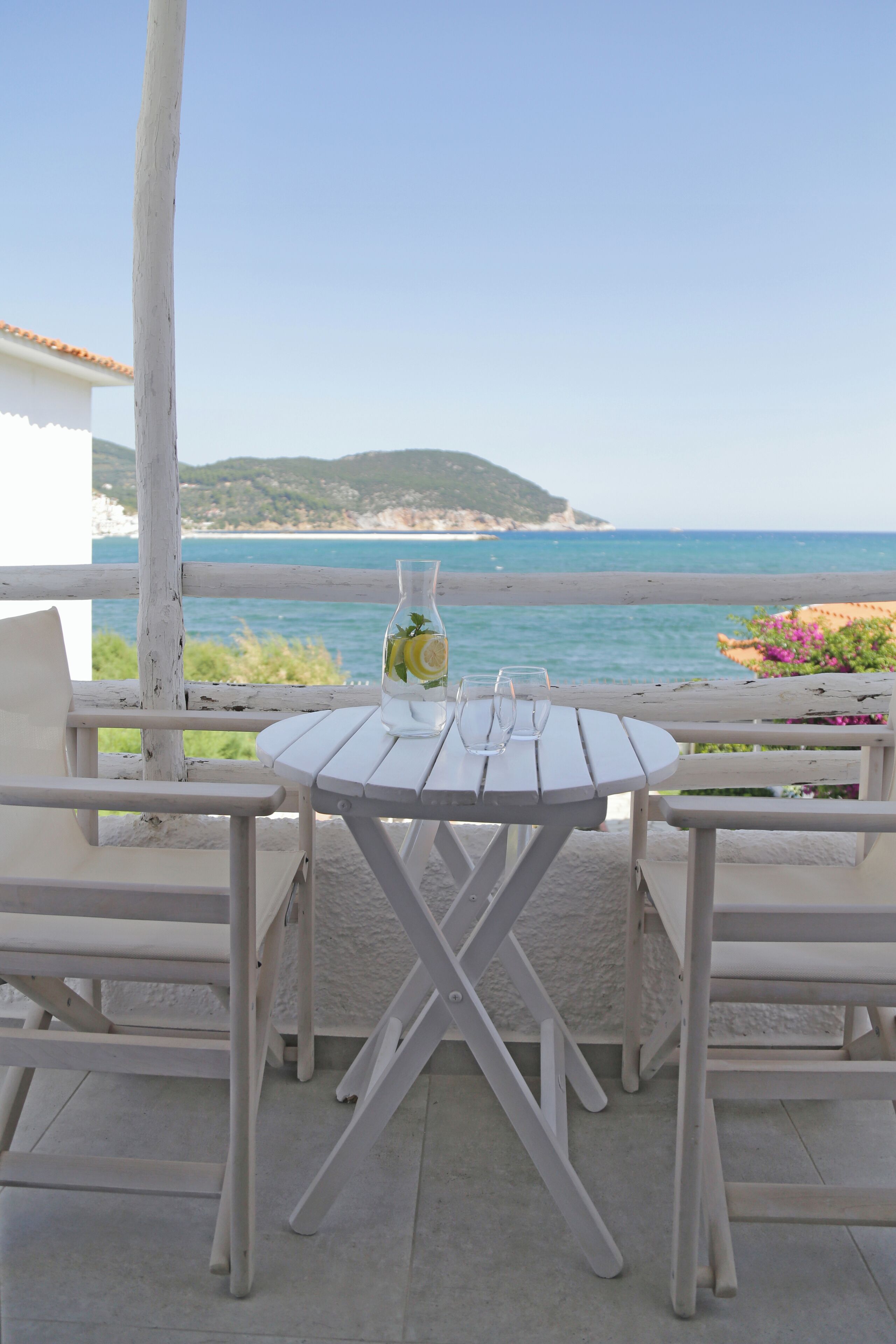 Une table en bois blanche, avec deux verres et une bouteille d’eau citronnée, est posée sur un balcon donnant sur une plage.