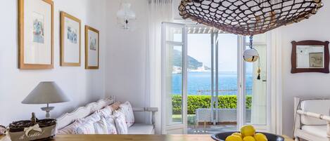 Una habitación con una mesa de madera, un cuenco de limones, cuadros en la pared y vista al mar a través de la puerta.
