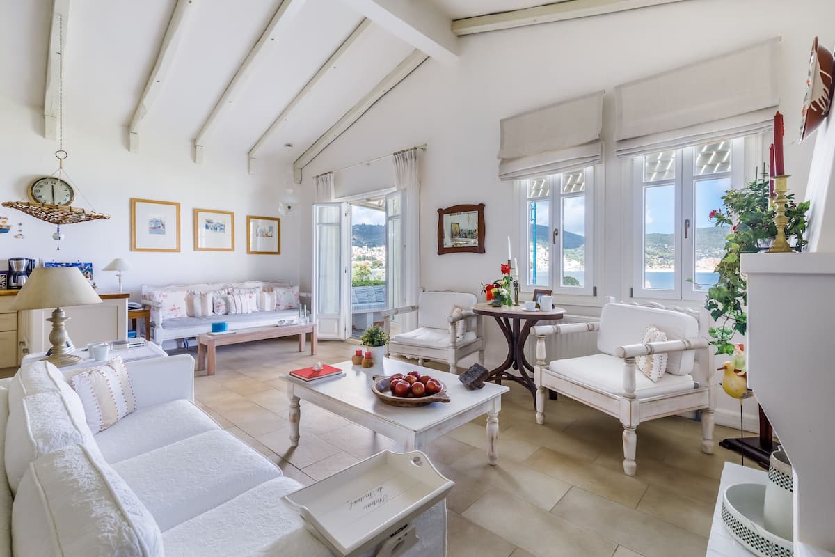 Un salón amplio con muebles blancos, una mesa de comedor con frutero, y ventanas grandes que ofrecen vista al mar.