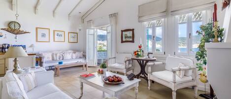 A spacious living room with white furniture, a dining table with a fruit bowl, and large windows offering a view of the sea.