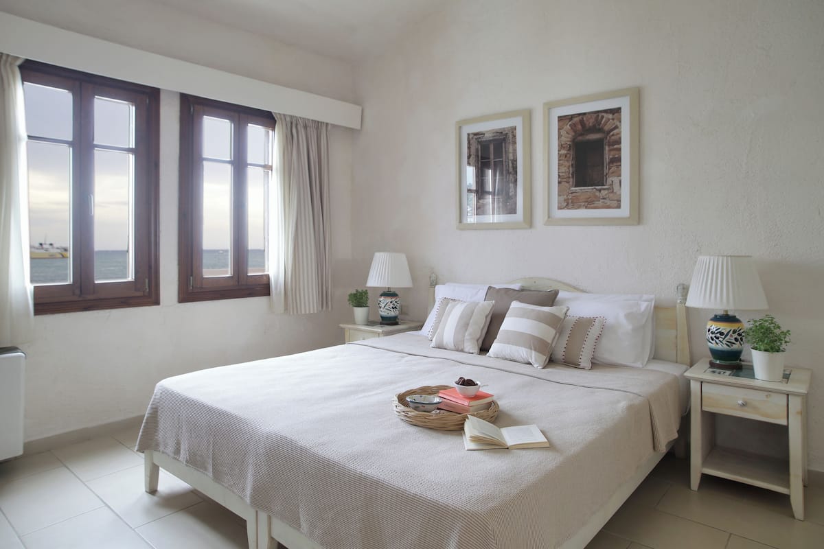 Un dormitorio ordenado con una cama grande, dos mesitas de noche, una ventana con cortinas y dos cuadros enmarcados en la pared.