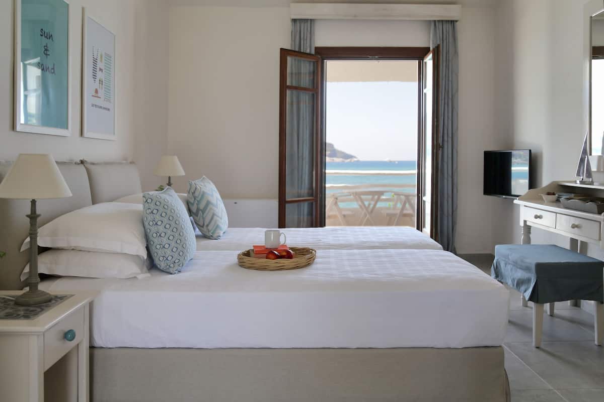 Habitación de hotel con cama, mesitas de noche, escritorio, televisor y vista a la playa.