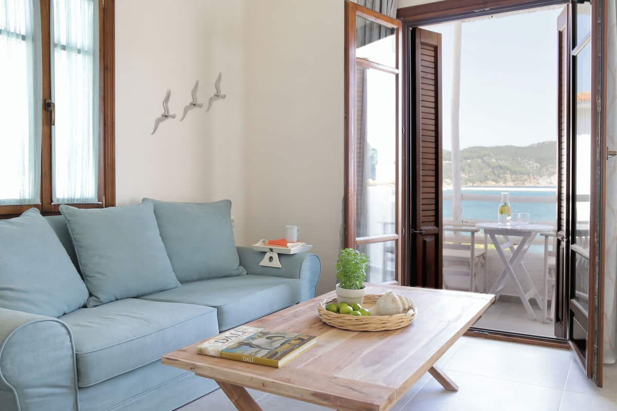 Una habitación con un sofá azul claro, una mesa de centro de madera con un libro y una cesta de fruta, y vista al mar a través de una puerta abierta.