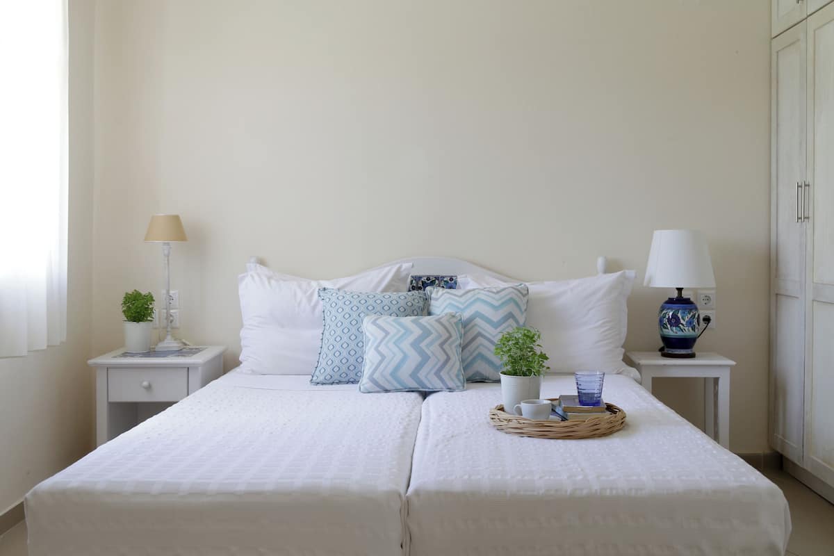 Una cama doble con ropa de cama blanca, dos mesitas de noche con lámparas, una pequeña planta en maceta y una bandeja de mimbre con una taza y un libro.