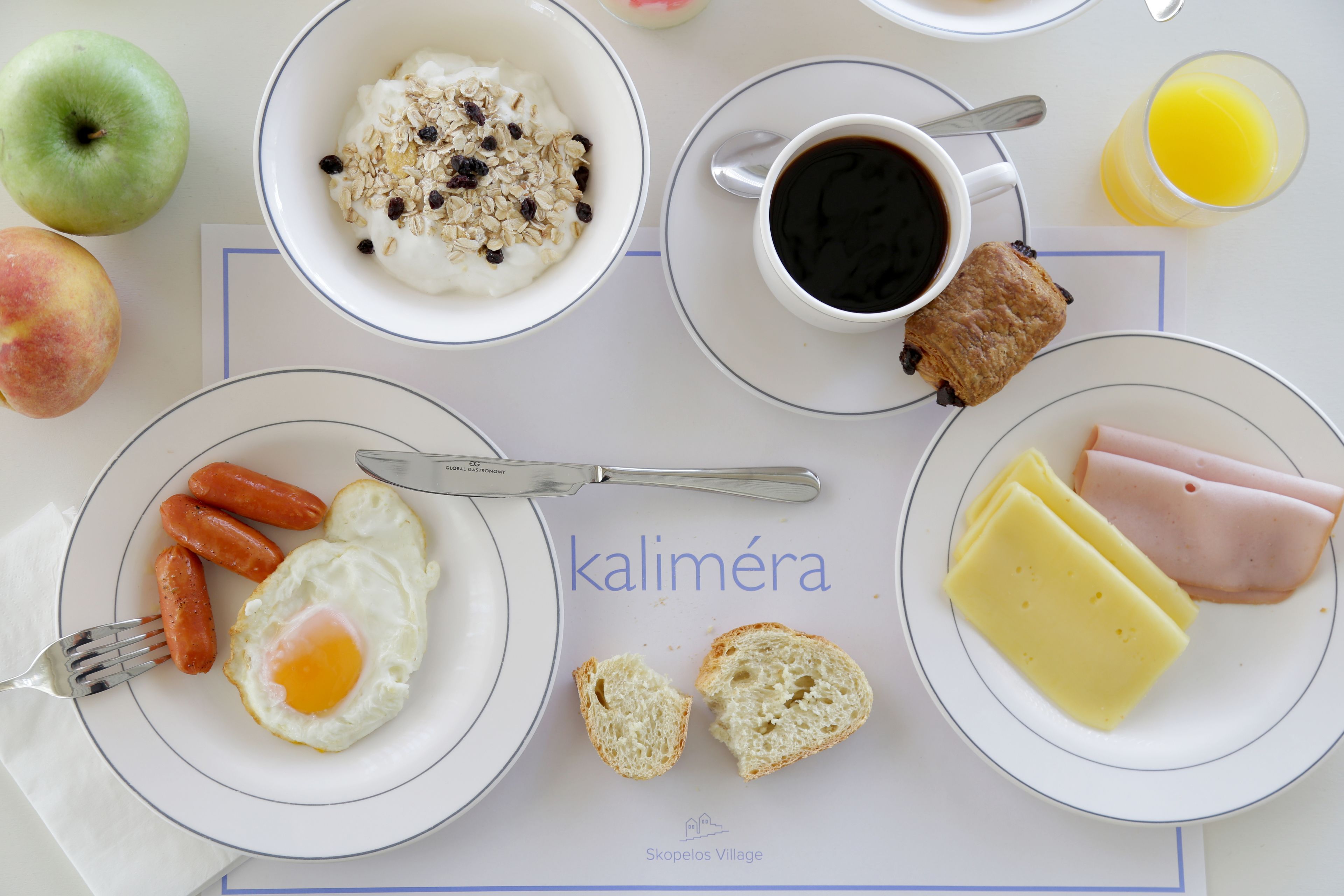 Una mesa de desayuno con platos de comida, incluyendo huevos, salchichas, jamón, queso y un tazón de yogur con fruta.