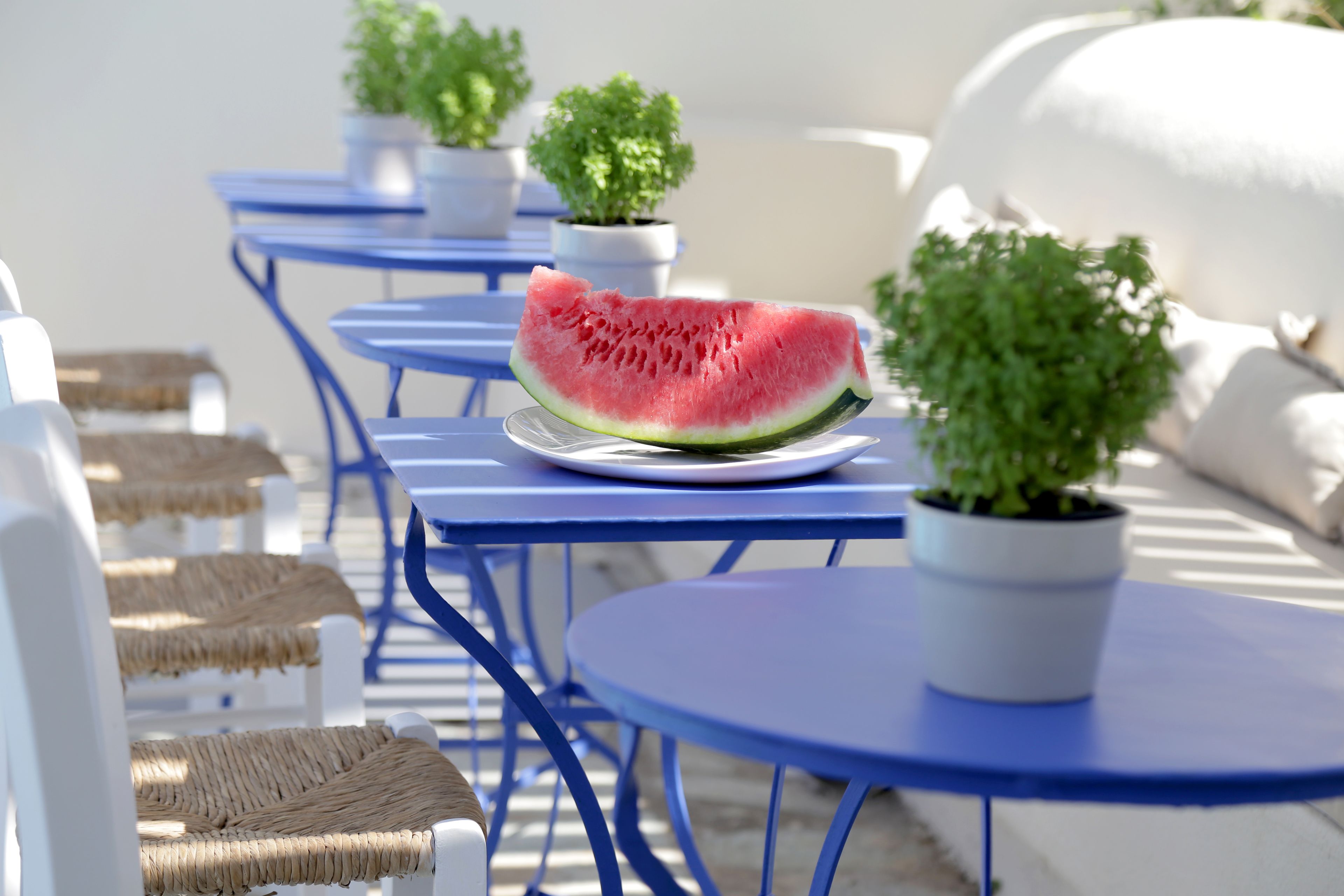 Eine Terrasse mit blauen Tischen und Stühlen, einem Teller mit Wassermelone und Topfpflanzen.