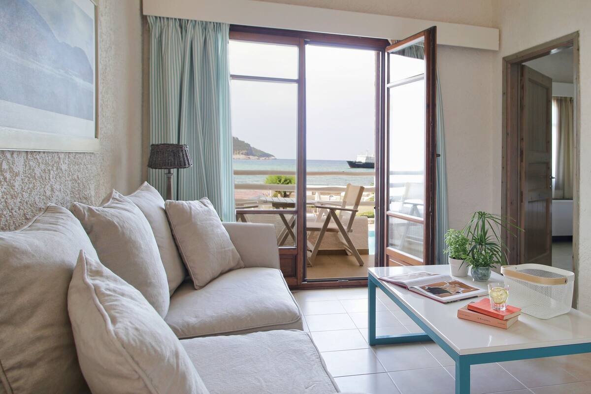 Una sala de estar con un sofá, una mesa con libros y un vaso, y vista al mar a través de un ventanal.