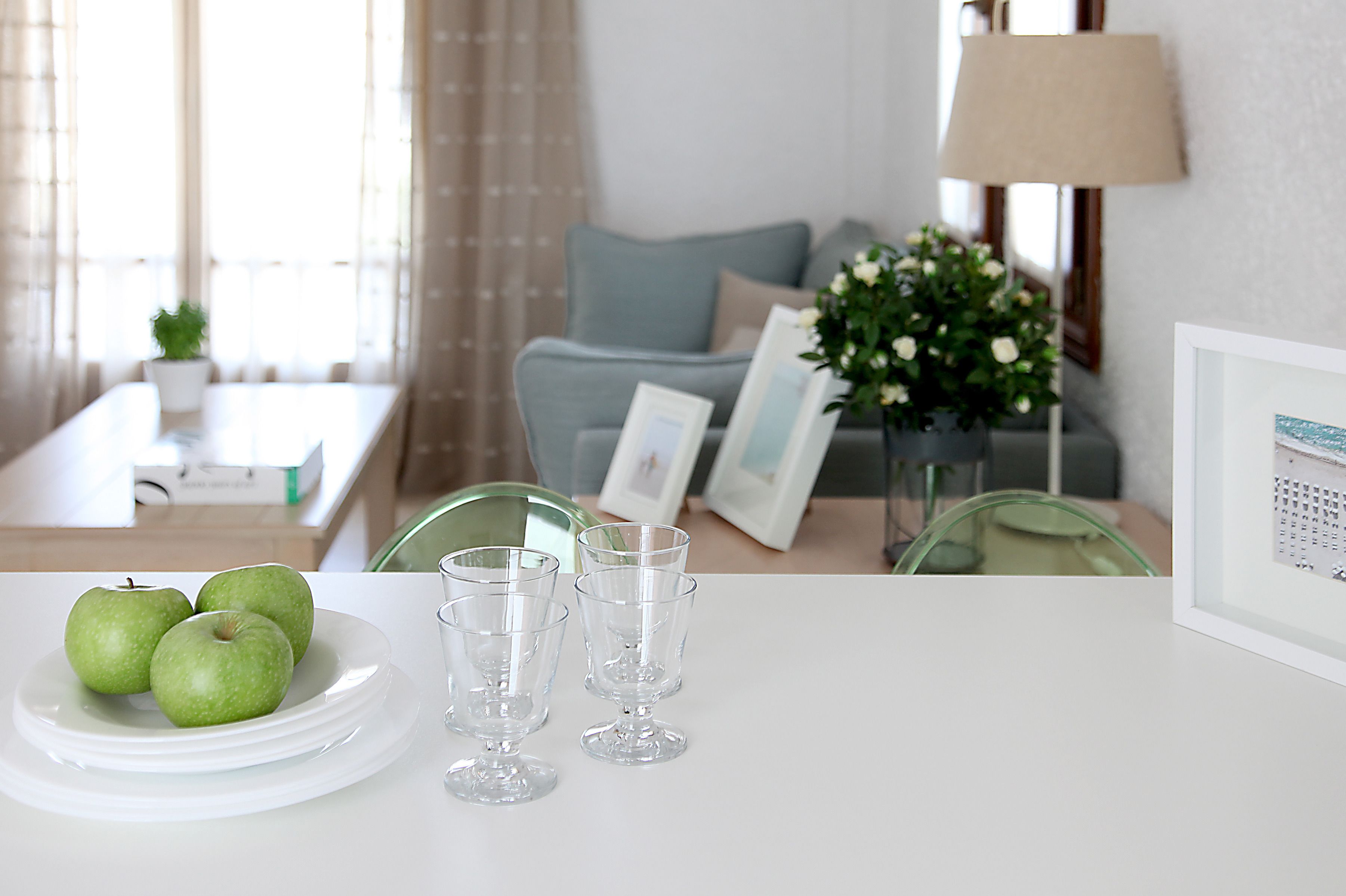 A dining table with a plate of green apples and three clear glasses, a green chair, and a framed picture on the table.