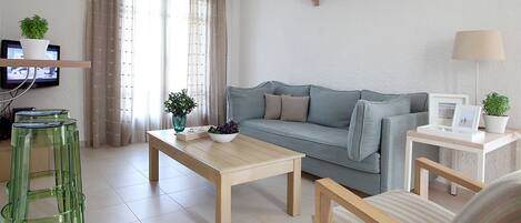 En stue med sofa, sofabord og to sideborde, et fjernsyn på et stativ og et vindue med gardiner.
