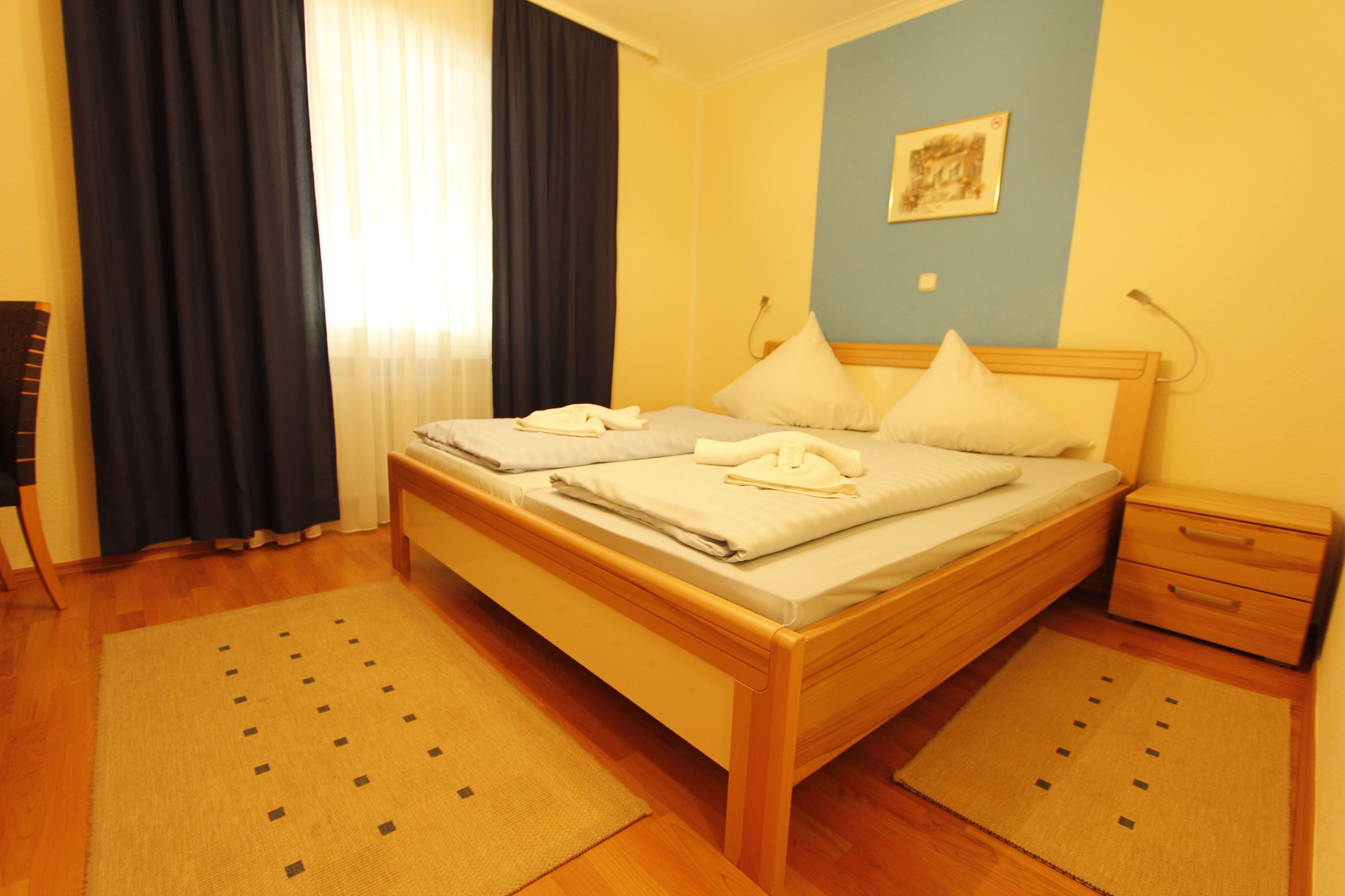 A hotel room with a wooden bed, two pillows, a nightstand, and a small drawer.
