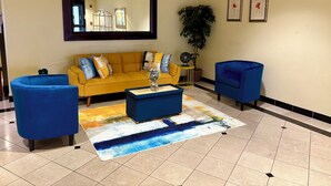 A lobby with a yellow sofa, blue armchairs, a coffee table, a mirror, and two framed pictures on the wall. - Days Inn & Suites by Wyndham Laurel Near Fort Meade (Laurel)