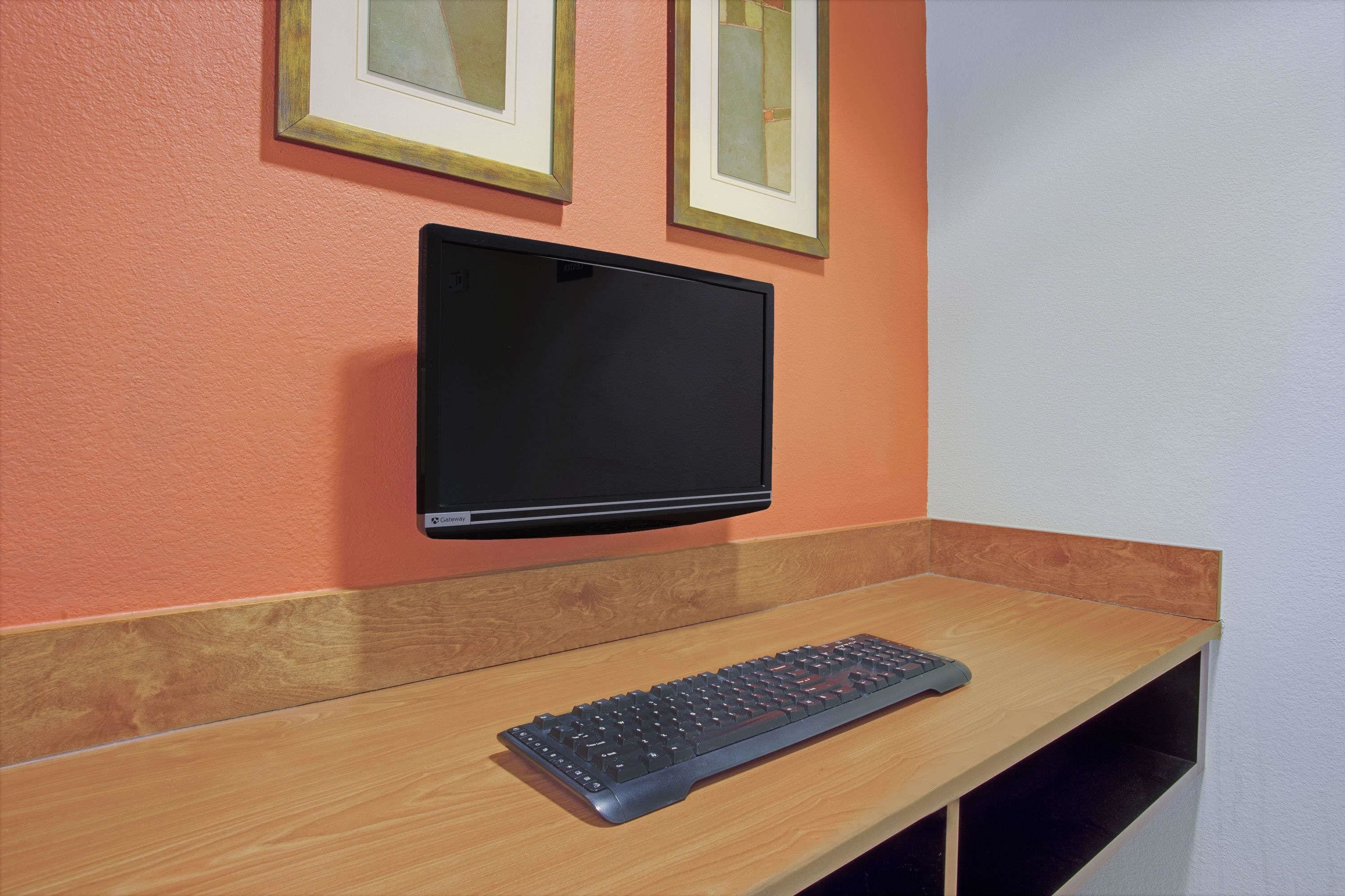 A computer monitor mounted on a wall above a wooden desk with a keyboard.