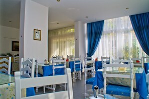 Uma área de refeições com cadeiras e mesas brancas, toalhas de mesa azuis e vista para o verde lá fora.