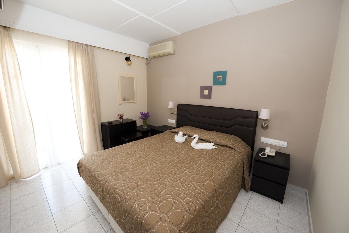 A hotel room with a large bed, a bedside table, a nightstand, a wall-mounted air conditioner, and a window with curtains.