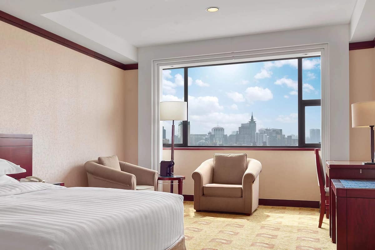 A hotel room with a large window, a bed, two armchairs, a desk, and a lamp.