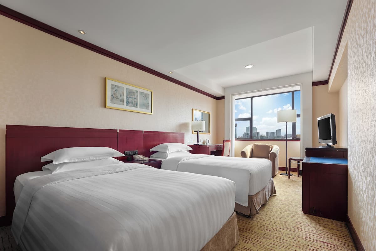 A hotel room with two beds, a desk with a television, a chair, and a large window with a city view.