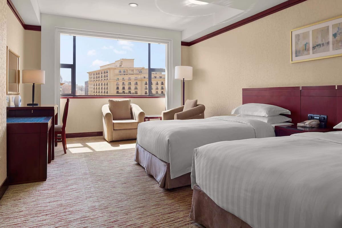 A hotel room with two beds, a desk, a chair, and a large window offering a city view.