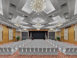A conference room with rows of white chairs, a large chandelier, and a stage with curtains.