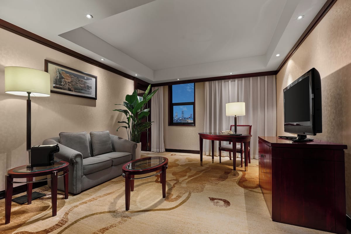 A hotel room with a sofa, a coffee table, a desk with a lamp, a television, and a large window with curtains.