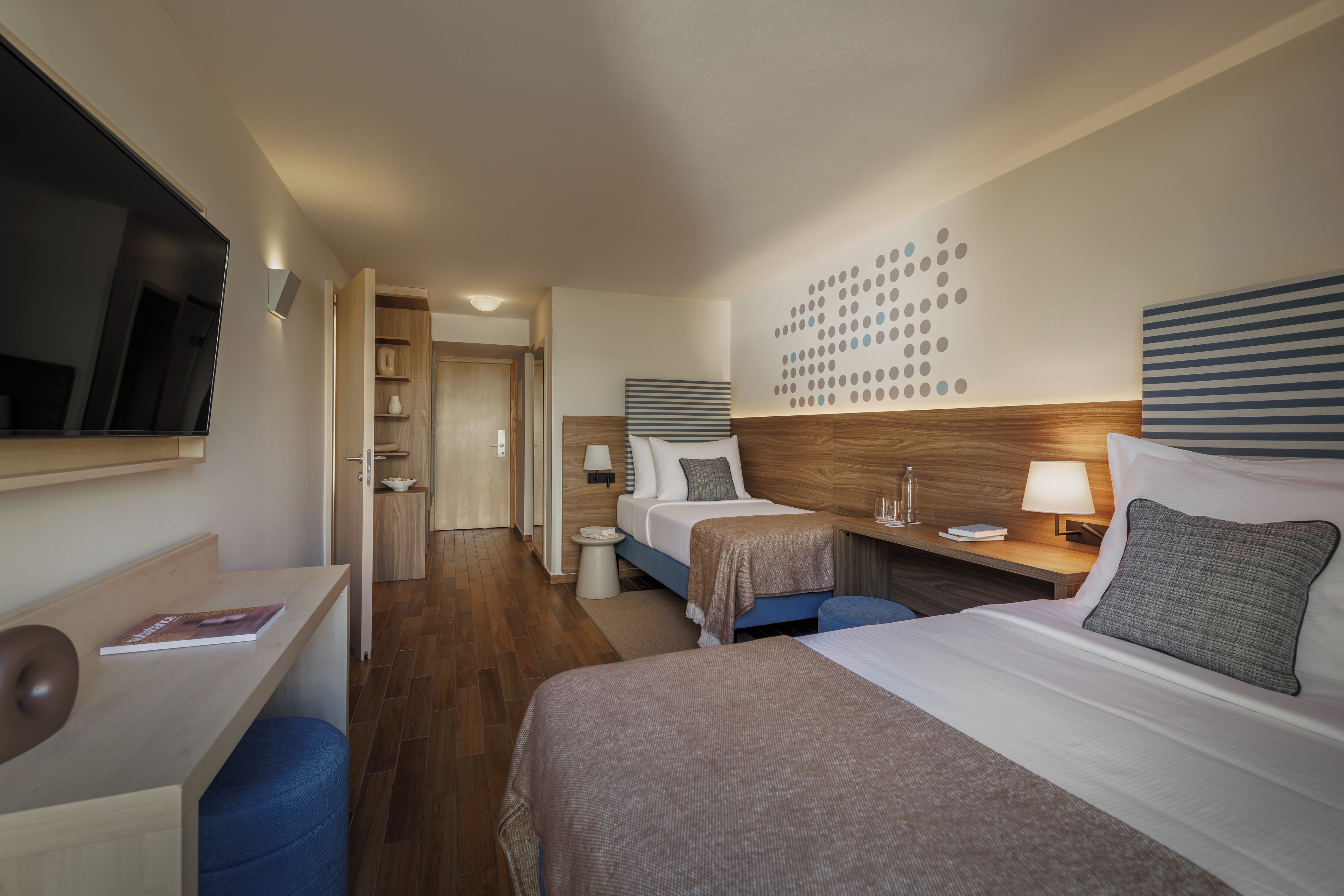 A hotel room with two beds, a desk, a chair, a bedside table with a lamp, and a wall with a circular pattern.