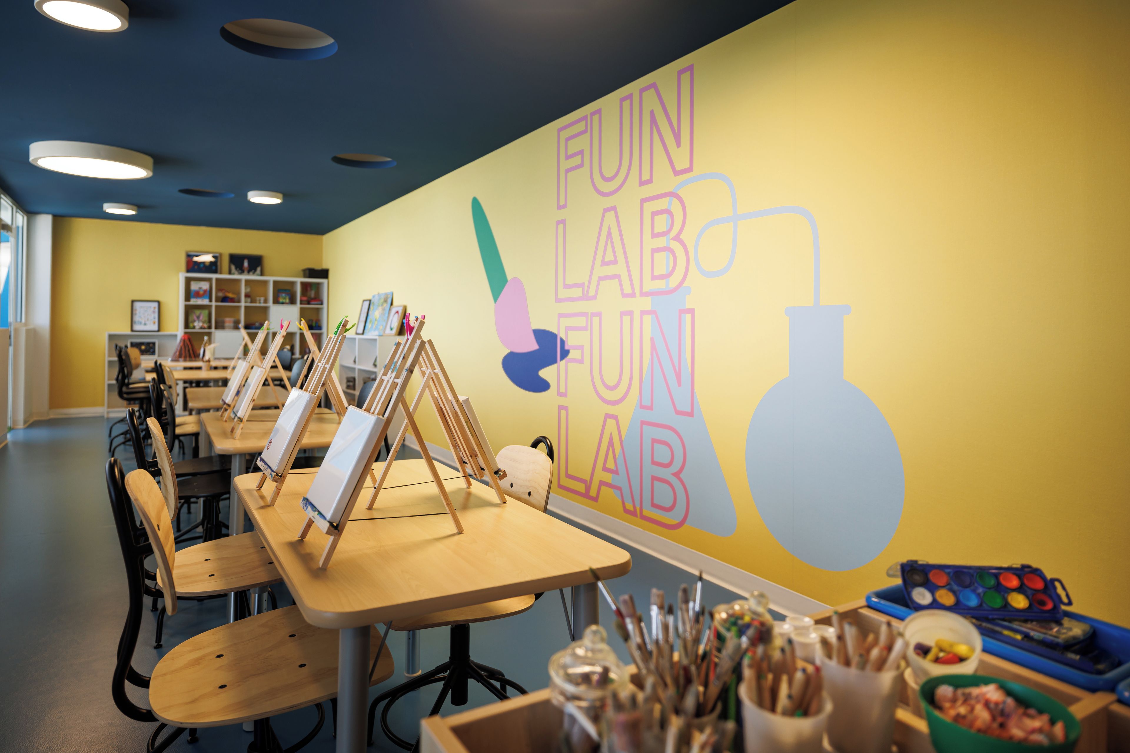 Un aula con mesas, sillas y material de arte, con una pared que muestra la inscripción 'FUN LAB FUN'.