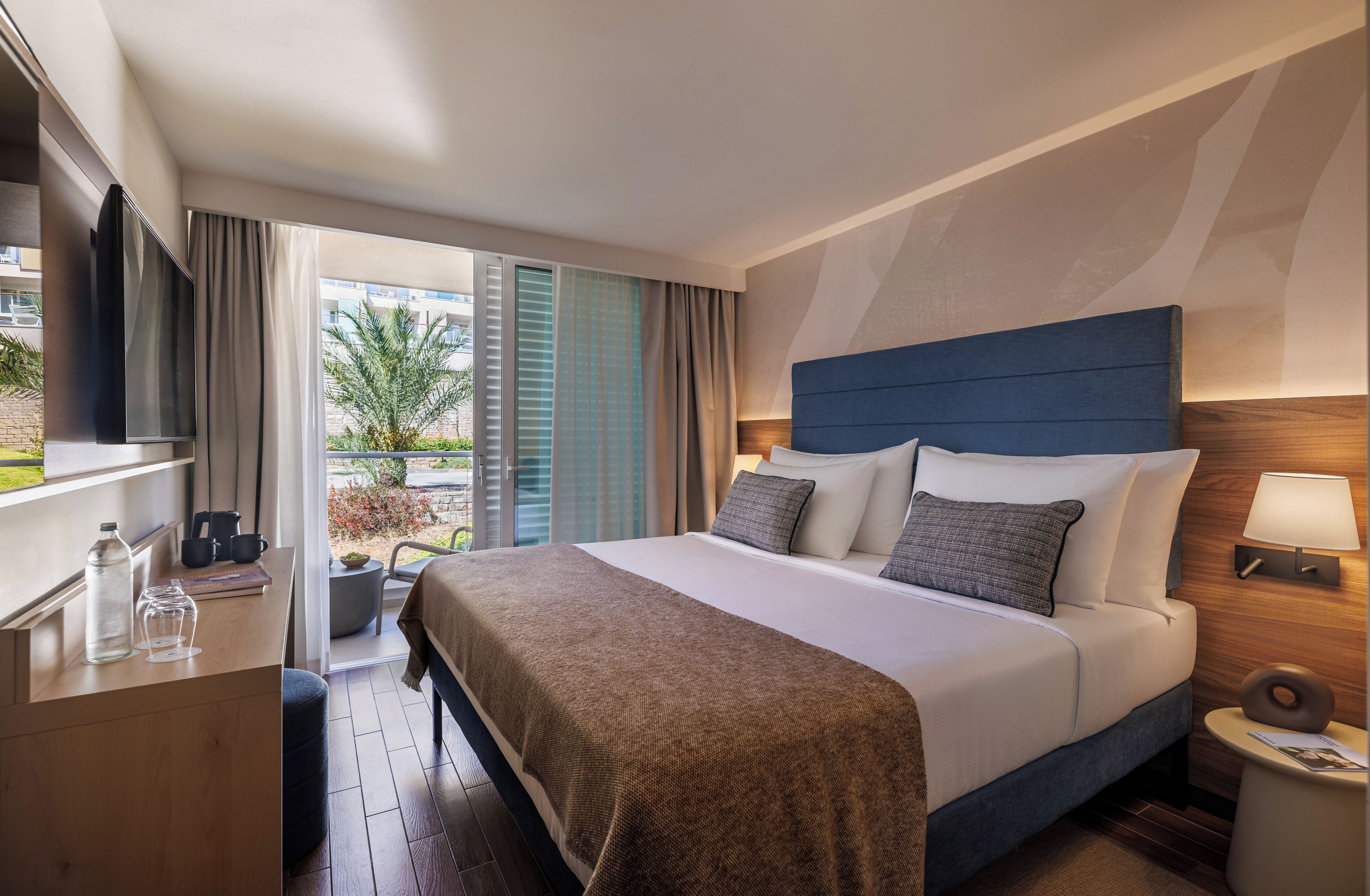 A hotel room with a bed, bedside table, lamp, and a view of the outdoors.