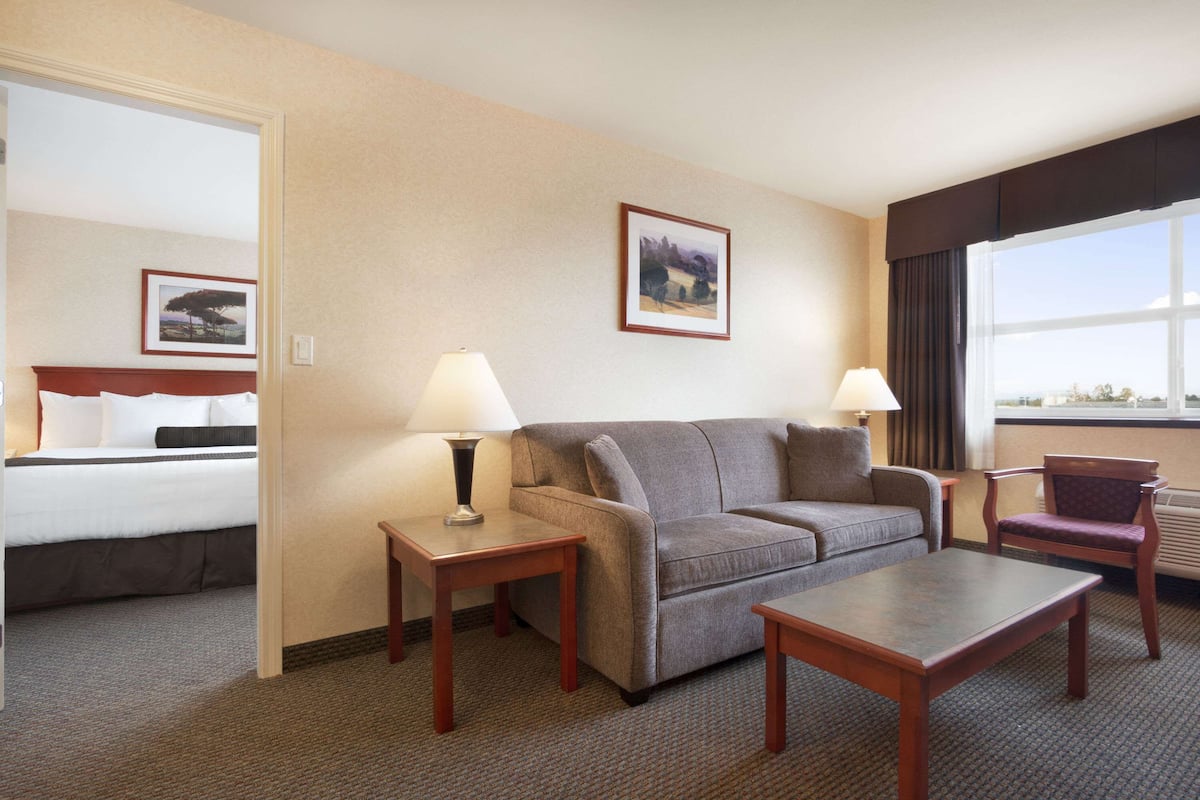 Un cuarto de hotel con una cama, un sofá gris, una mesa de centro de madera, dos mesitas de noche con lámparas, una silla y cuadros enmarcados en la pared.