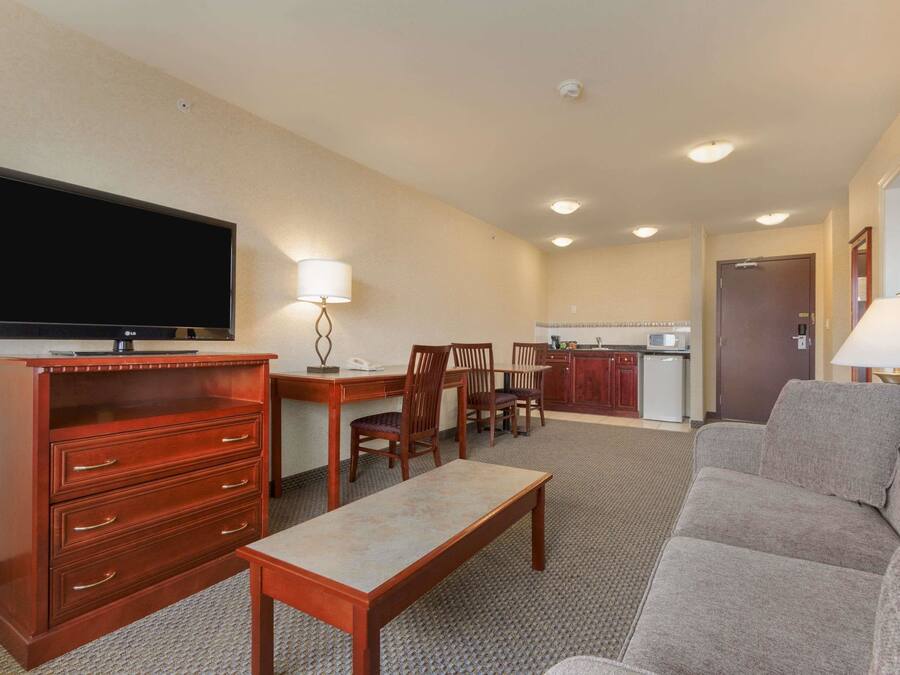 A hotel room with a flat-screen TV, wooden furniture, a sofa, a dining table, and a kitchenette.