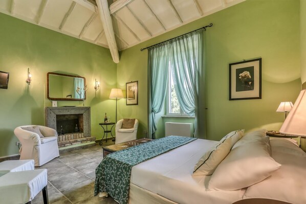 A room with a bed, two chairs, a fireplace, a table, and a window with curtains. - Relais Pian di Vico- Guest House (Tuscania)