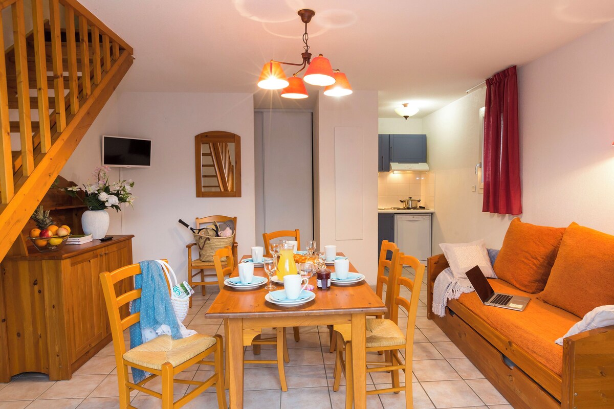 A cozy living room with a wooden staircase, a dining table set for four, and a sofa with a laptop.