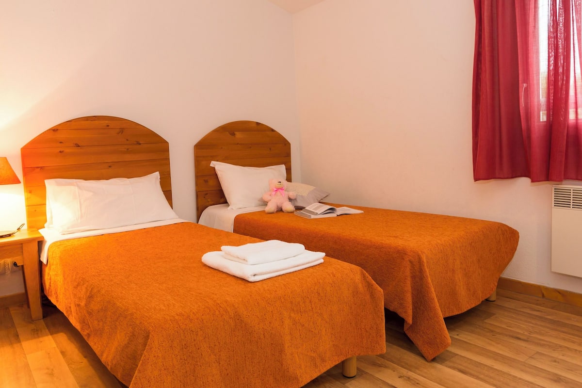Una habitación con dos camas, una mesita de noche, una lámpara, un peluche de oso y una ventana con cortinas rojas.