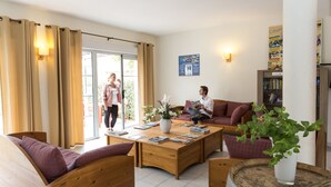 A cozy living room with wooden furniture, a coffee table with books, a bookshelf, and a large window with curtains. - Odalys Residence Coeur d’Ardèche (Vallon-Pont-d'Arc)