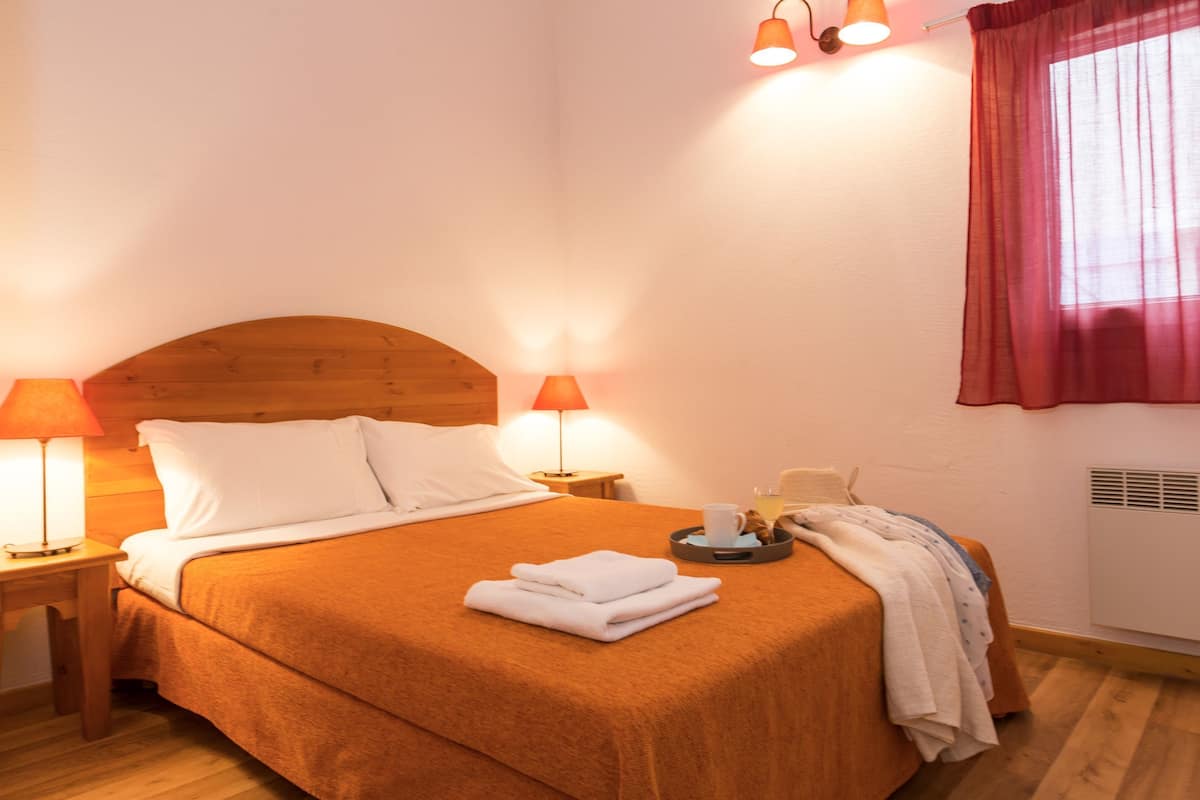 Una cama bien hecha con cabecera de madera, dos lámparas de noche, una bandeja con una taza y pasteles, y una ventana con cortina roja.