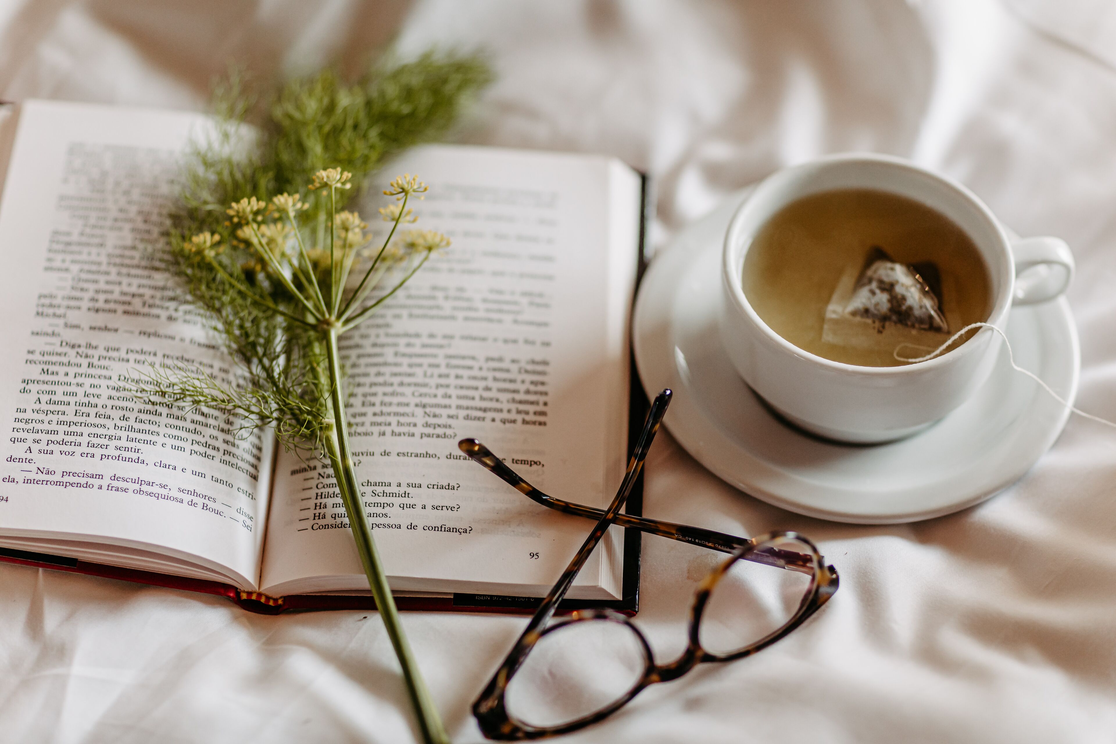 Un livre ouvert, une tasse de thé et une paire de lunettes posés sur une surface blanche.