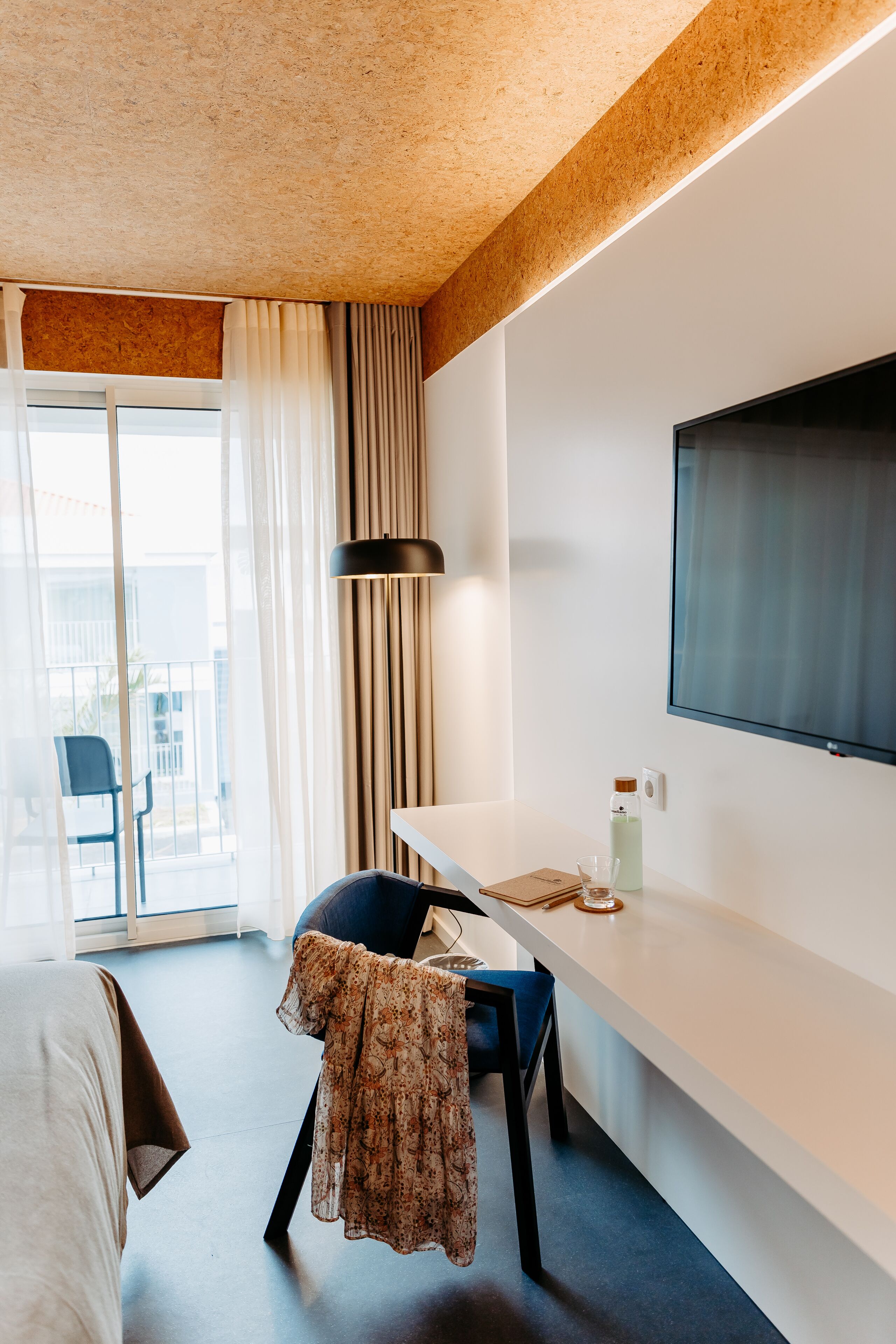 A modern hotel room with a large window, a flat-screen TV mounted on the wall, a desk with a chair, and a bed.