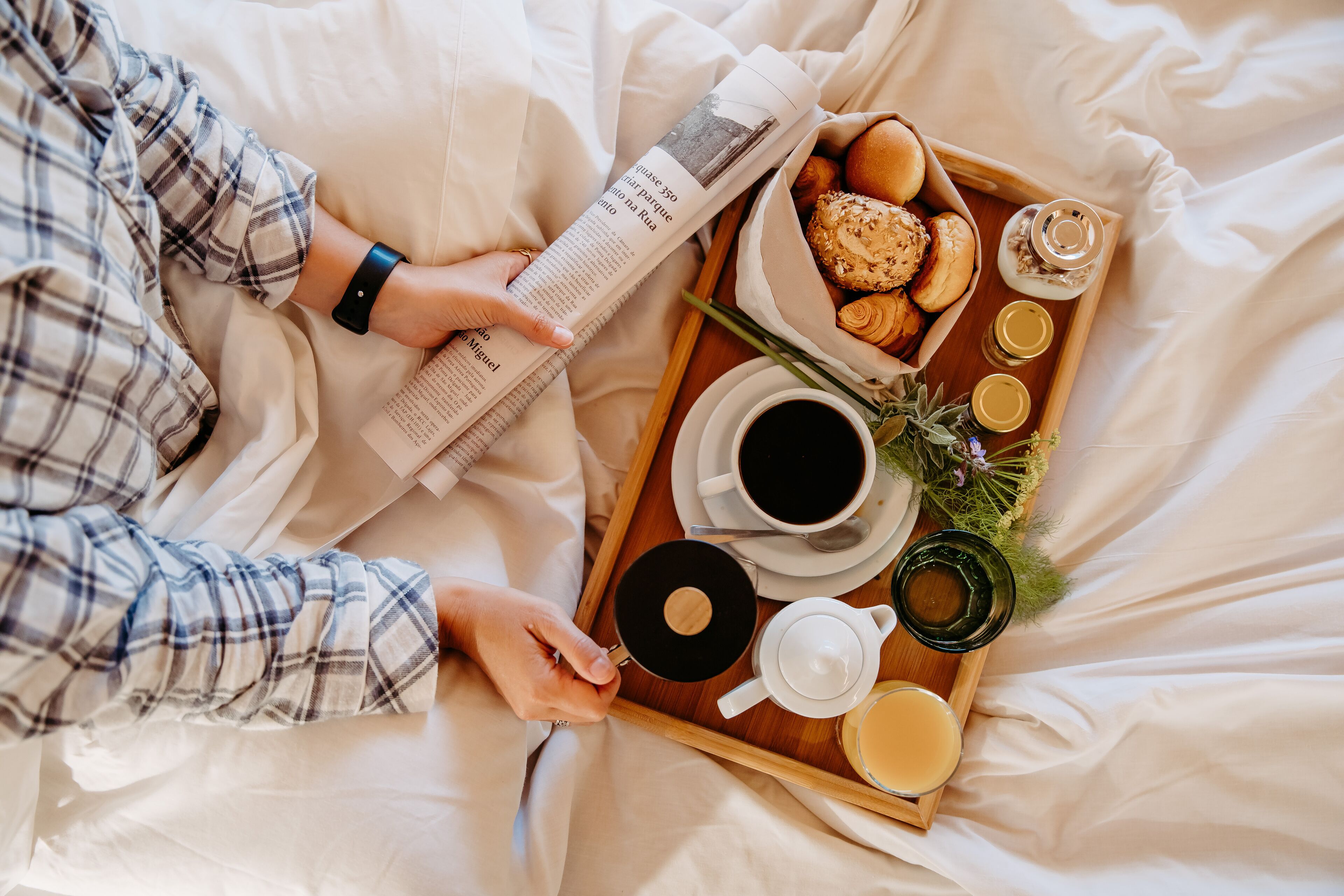 A person reading a newspaper on a bed with a tray containing food and drinks.