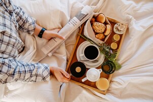 Una persona leyendo un periódico en una cama, con una bandeja que contiene comida y bebidas.