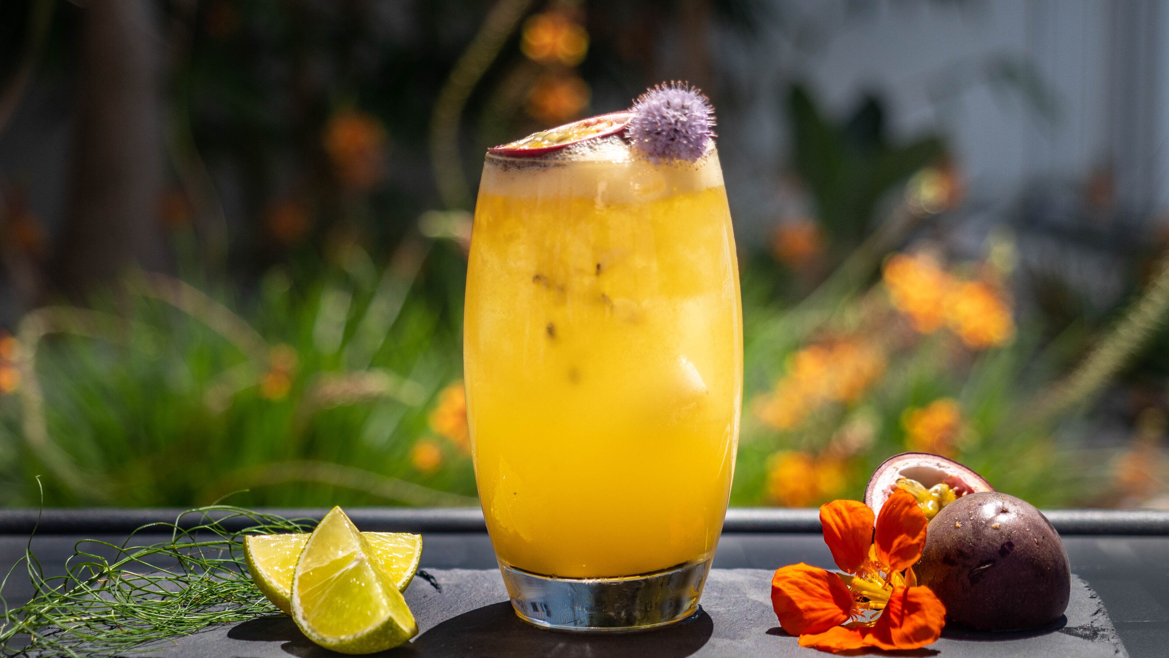 Un vaso alto de jugo de naranja con una rodaja de lima y una flor como adorno, sobre una mesa con superficie oscura.