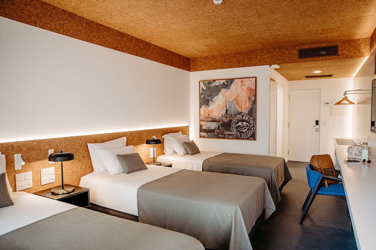 A hotel room with two beds, a desk, a chair, a coat rack, and a painting on the wall.