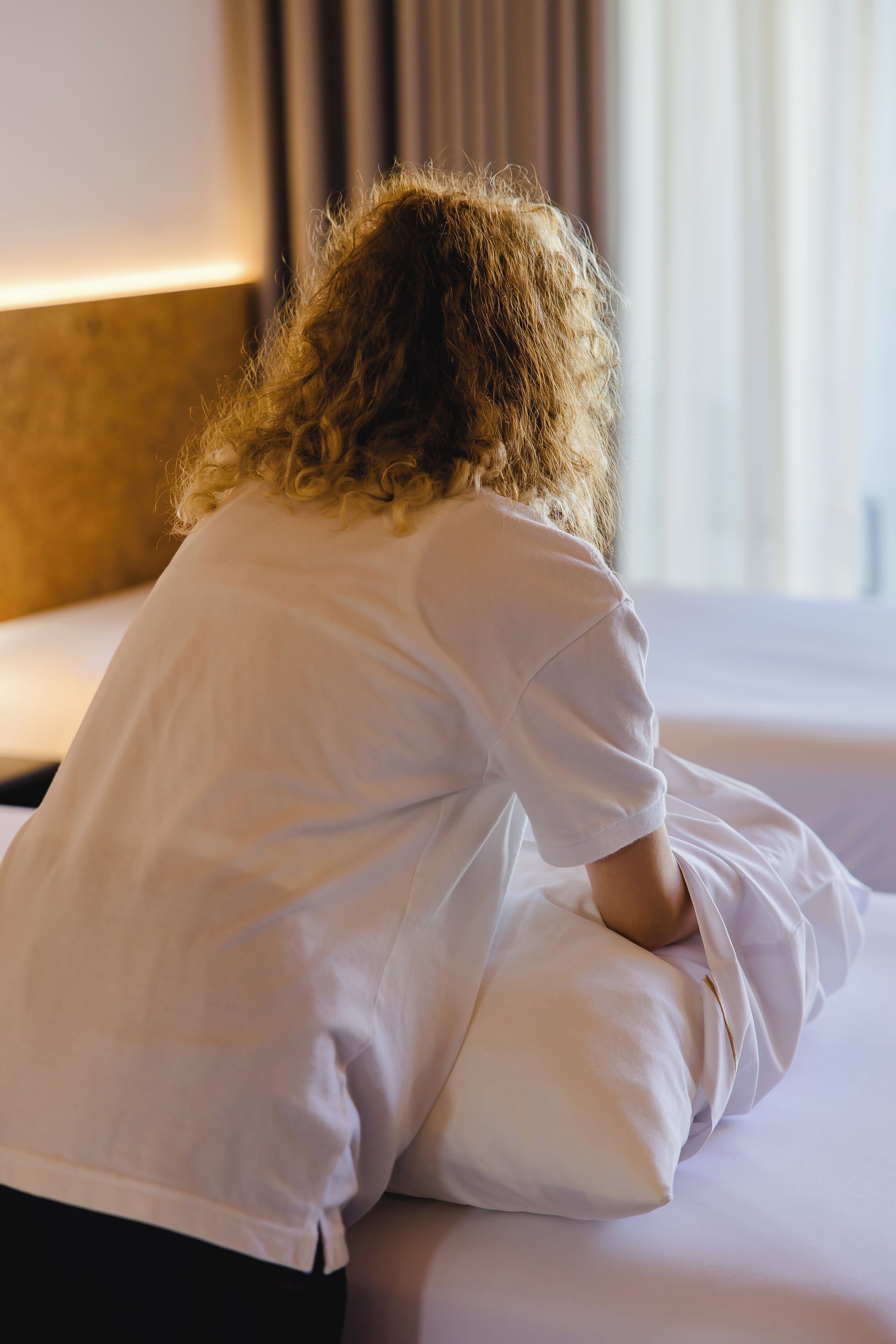 A person sitting on a bed in a room with a headboard and a window with curtains.