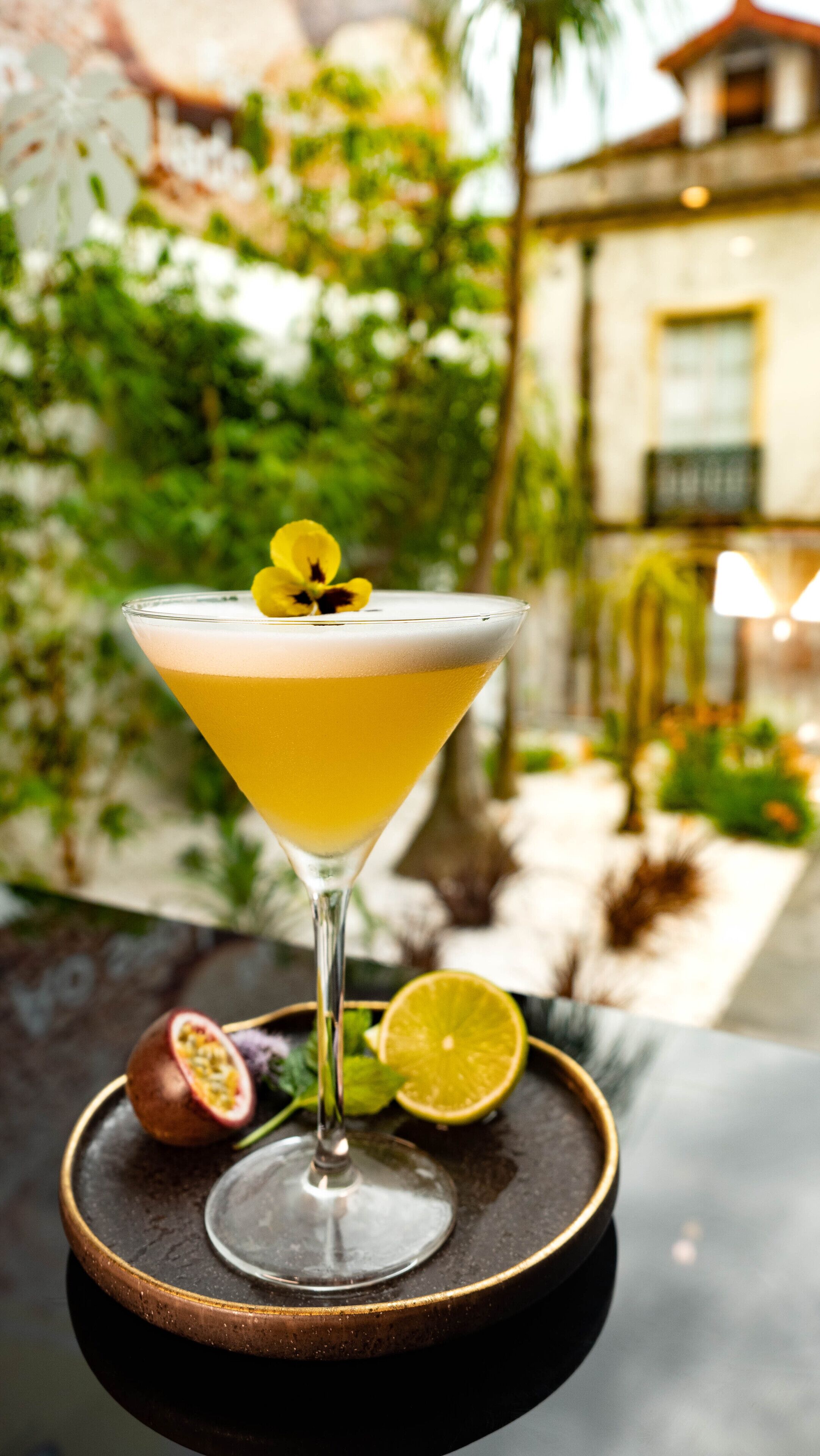 A cocktail in a martini glass with a yellow garnish, placed on a tray with a slice of lime and a passion fruit.